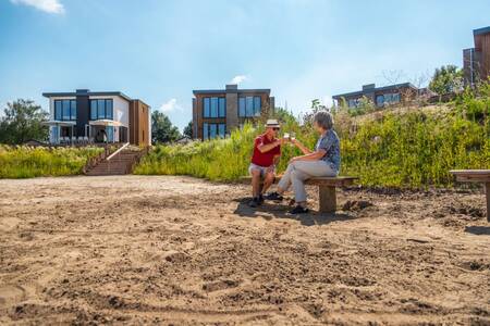 A couple is sitting on a bench on the beach of the EuroParcs Aan de Maas holiday park